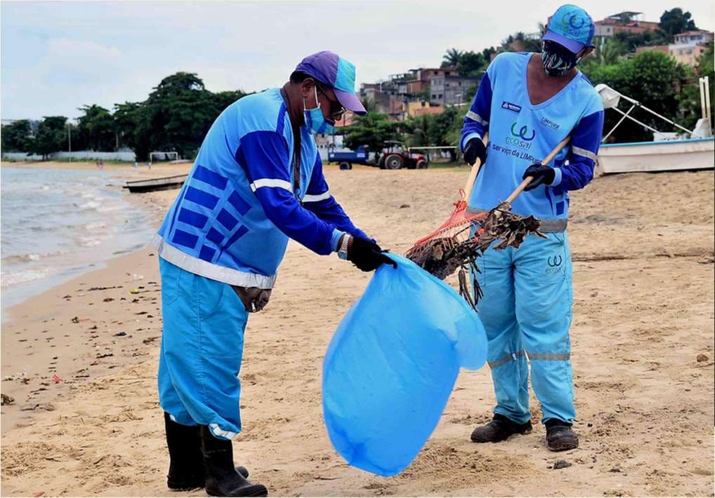 Dois agentes de limpeza de praia recolhendo resíduos da areia com rodo e saco